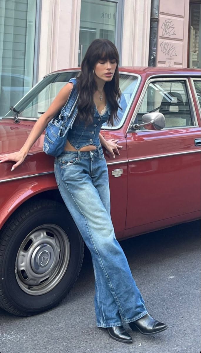 Woman in a denim vest and jeans leaning on a red car on a street