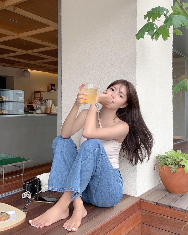 Woman with long hair sitting barefoot outdoors, holding a drink