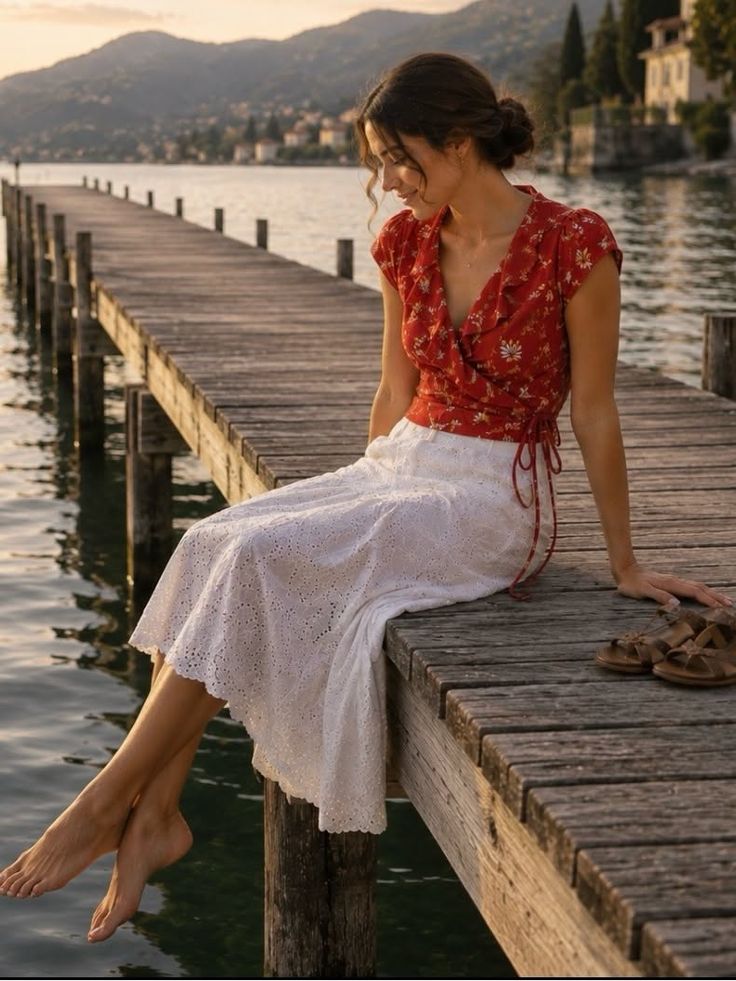Woman in a red floral top and white skirt sitting on a wooden pier at golden hour