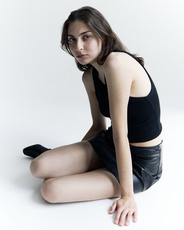 Woman in a black tank top and leather skirt sitting on a white background