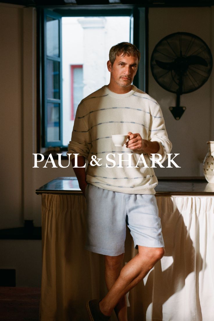 Man in a striped sweater and light blue shorts holding a cup indoors