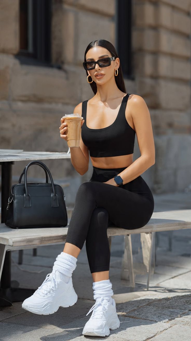 Woman in black activewear and sunglasses sitting outdoors with an iced coffee
