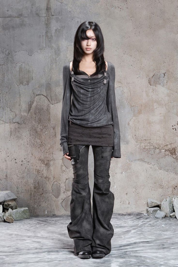 Woman with long black hair wearing dark, distressed clothing against a concrete wall