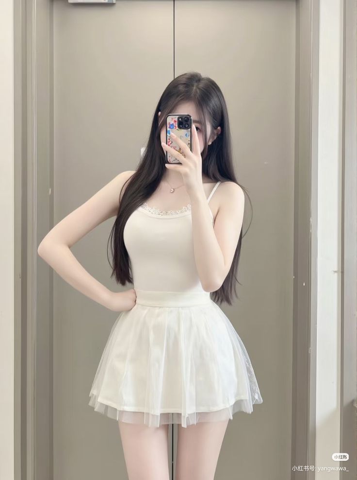 Woman in a white camisole and mini skirt taking a mirror selfie
