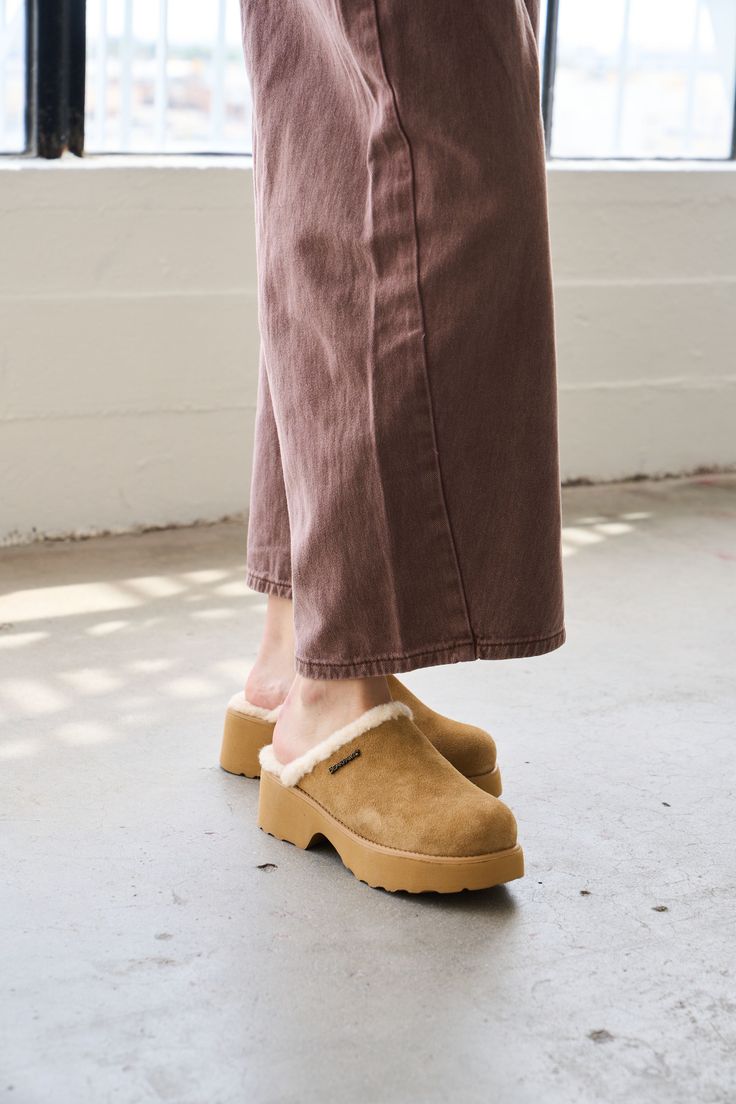 Person wearing brown pants and suede clogs with shearling lining on a concrete floor