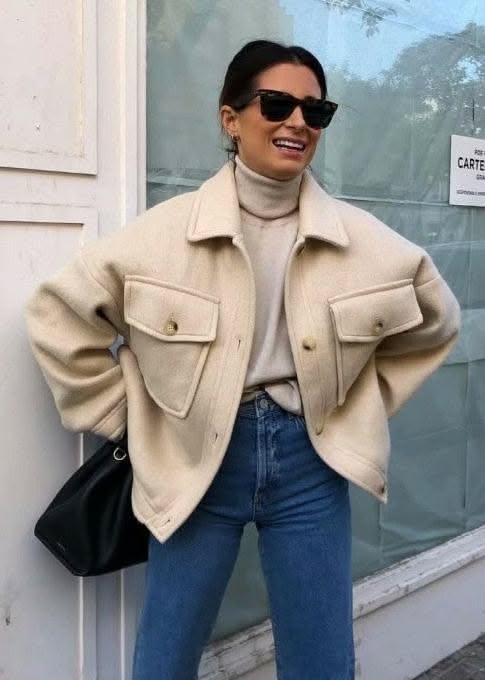Woman in a cream jacket, turtleneck, and jeans, smiling outdoors