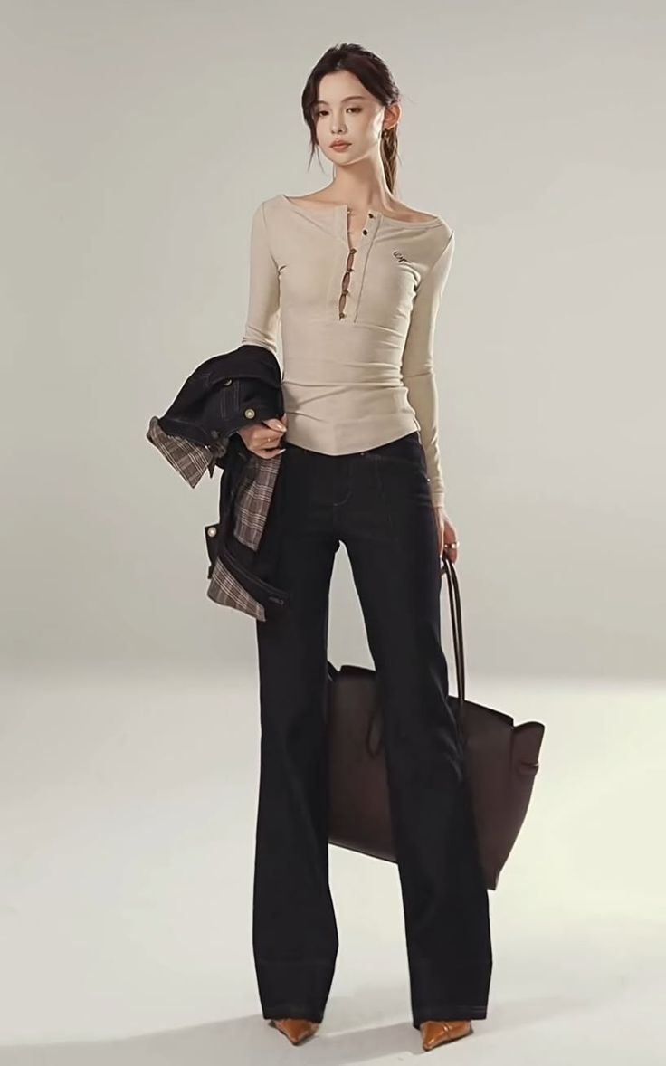 Woman in a long sleeve shirt and dark jeans holding a jacket and a bag