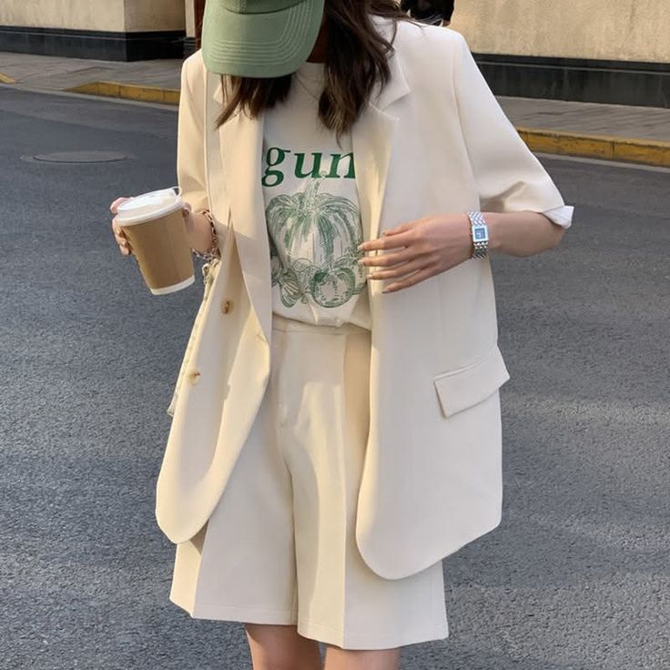 Person in a blazer, shorts, and baseball cap holding a coffee cup on a street