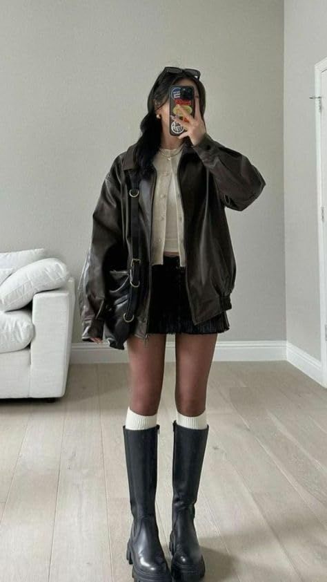 Person in a leather jacket, black skirt, and knee-high boots taking a mirror selfie
