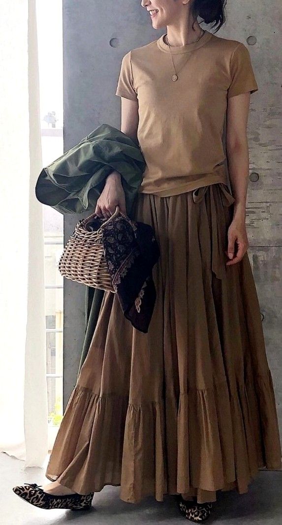 Woman in a brown top and long skirt holding a wicker bag and green jacket