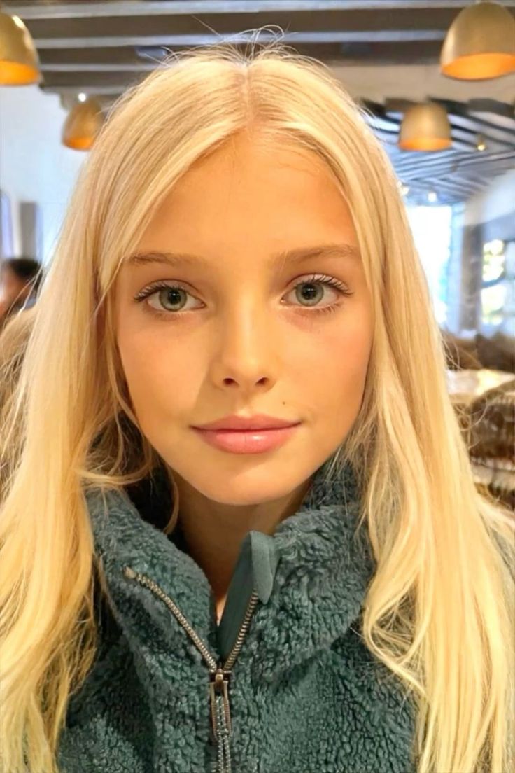 Young girl with blonde hair and blue eyes wearing a teal jacket indoors