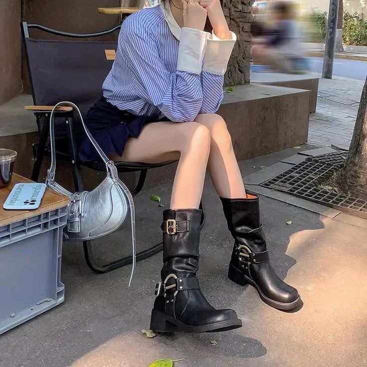 Person in a striped shirt and mini skirt sitting outdoors with black boots and a silver bag