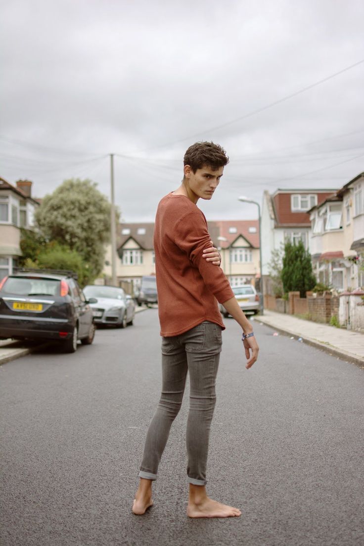 Young man in a rust-colored shirt and grey jeans standing barefoot on a street