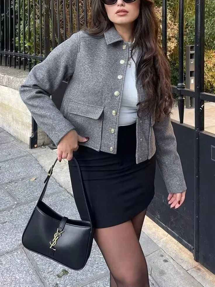 Woman in a gray jacket and black mini skirt holding a black handbag outdoors