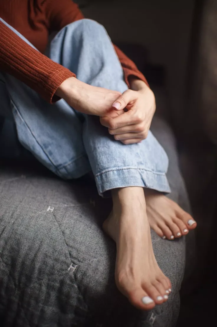 Person in blue jeans and a rust-colored top with bare feet on a gray blanket