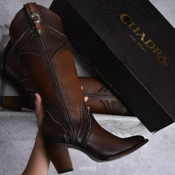 Brown leather cowboy boots with embossed details and a buckle, held by a hand