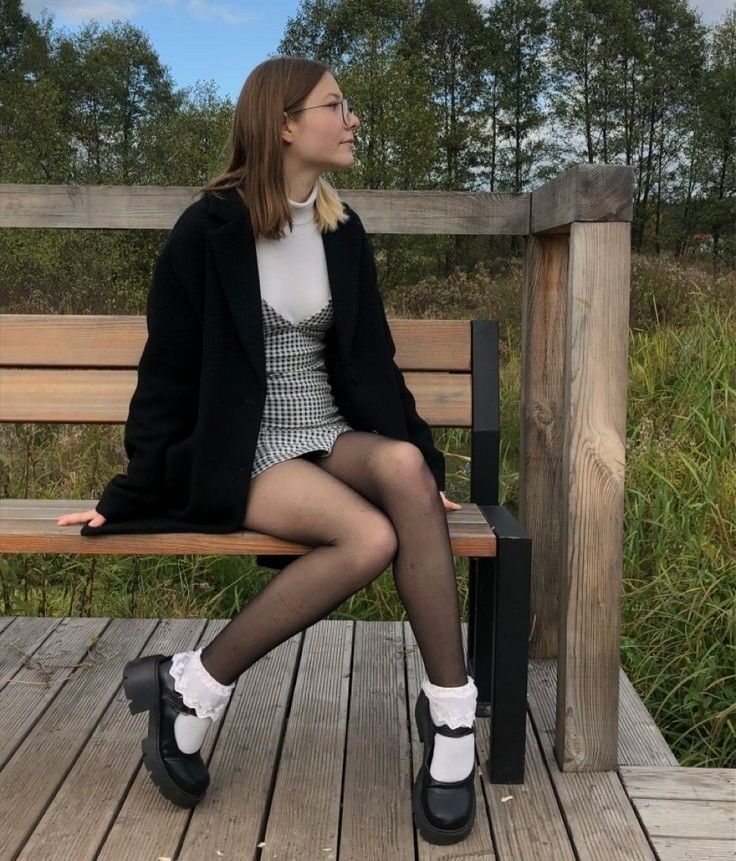 Woman in a black coat and gingham dress sitting on a wooden bench outdoors