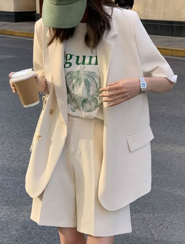 Person in a blazer, shorts, and baseball cap holding a coffee cup outdoors