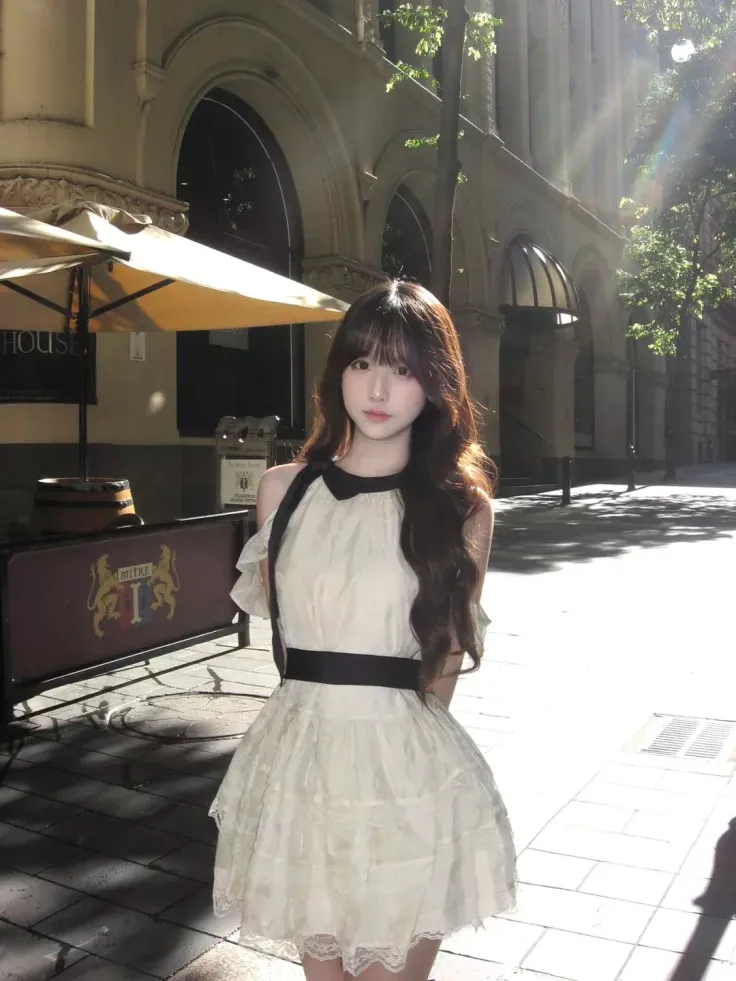 Woman in a white dress with a black collar and belt standing on a sunny street
