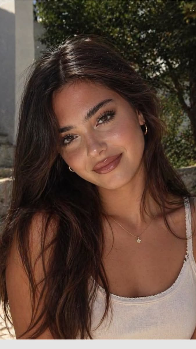 Woman with long dark hair wearing a white tank top and gold jewelry outdoors