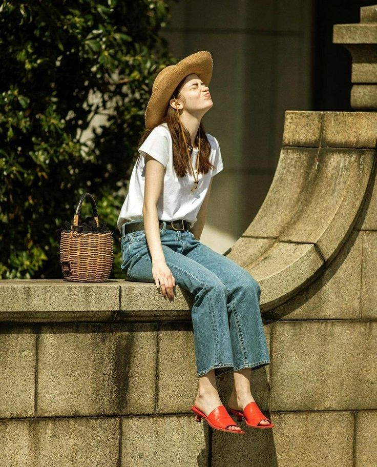 Woman in a straw hat and blue jeans sitting on a stone wall in bright sunlight