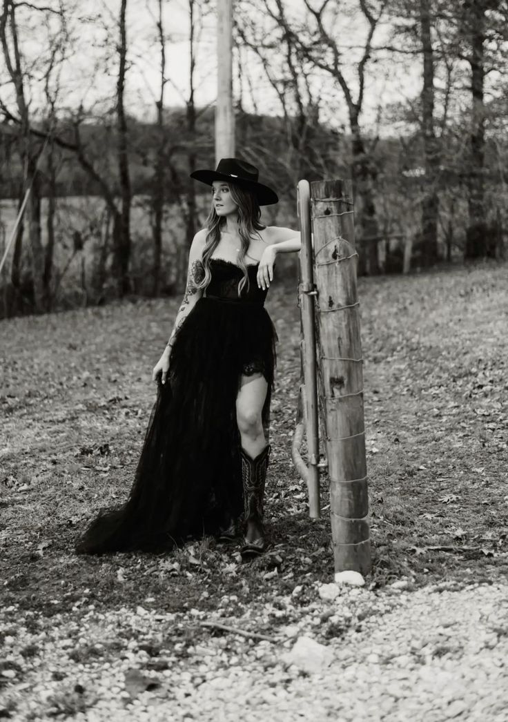 Woman in a black dress, cowboy hat, and boots leaning on a fence post in a forest