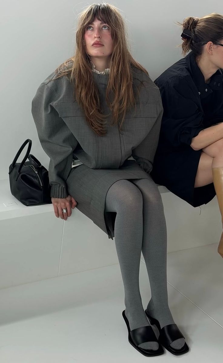 Woman with long hair in a gray outfit and tights sitting indoors