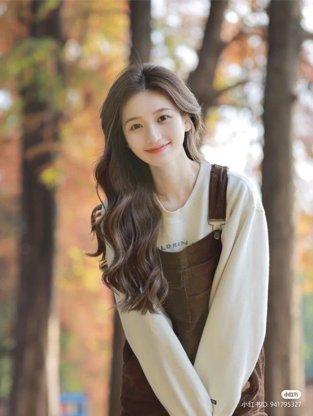 Woman with long wavy hair wearing brown overalls and a white long sleeve shirt outdoors