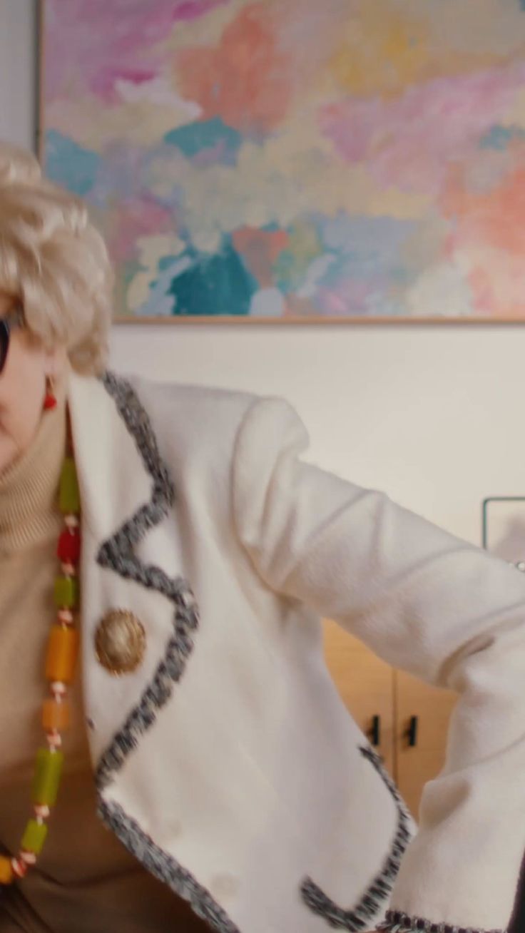 Person with blonde hair wearing a white jacket and colorful necklace indoors