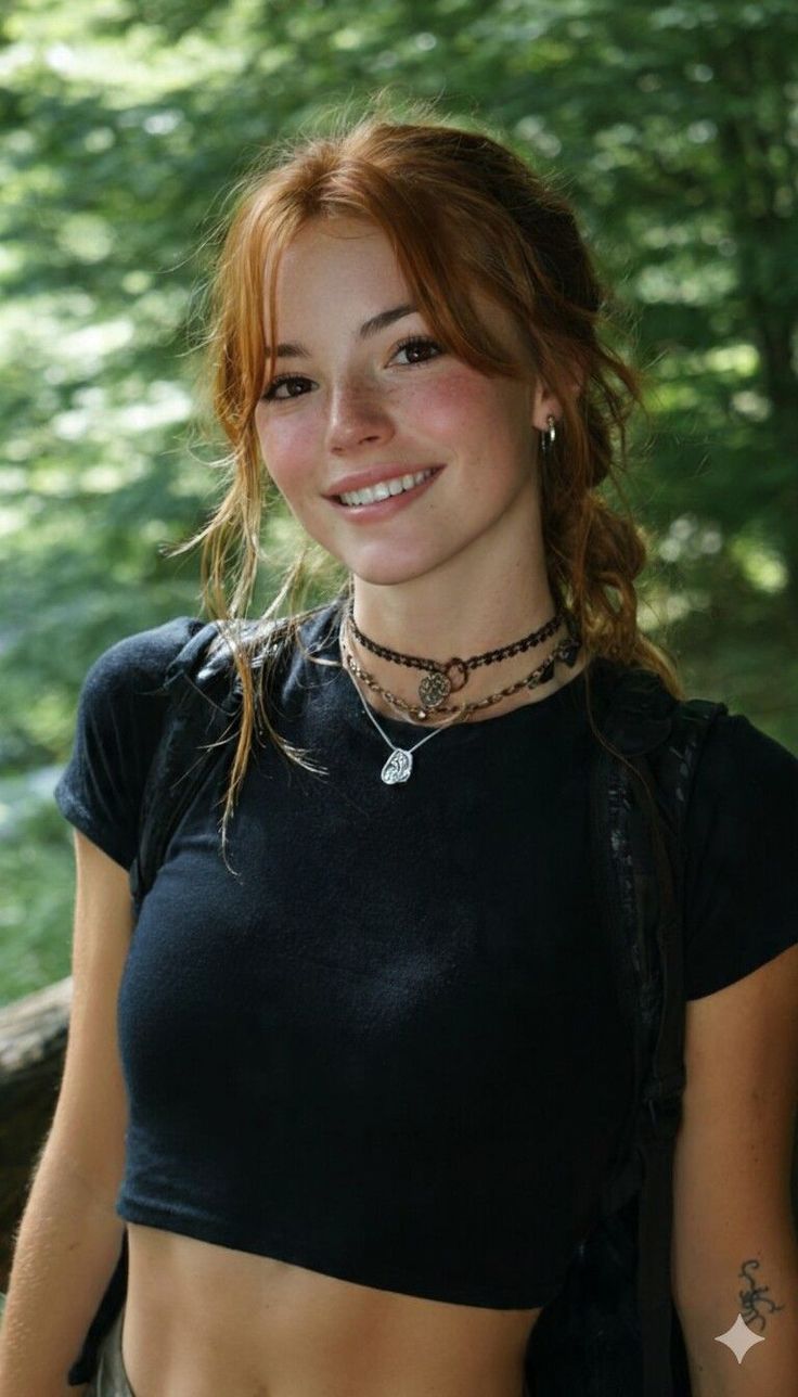Smiling woman with red hair and freckles wearing a crop top and layered necklaces outdoors