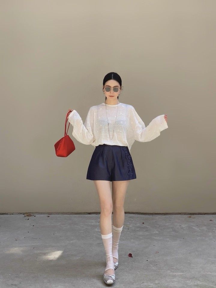Woman in sunglasses and a white top holding a red handbag, standing outdoors