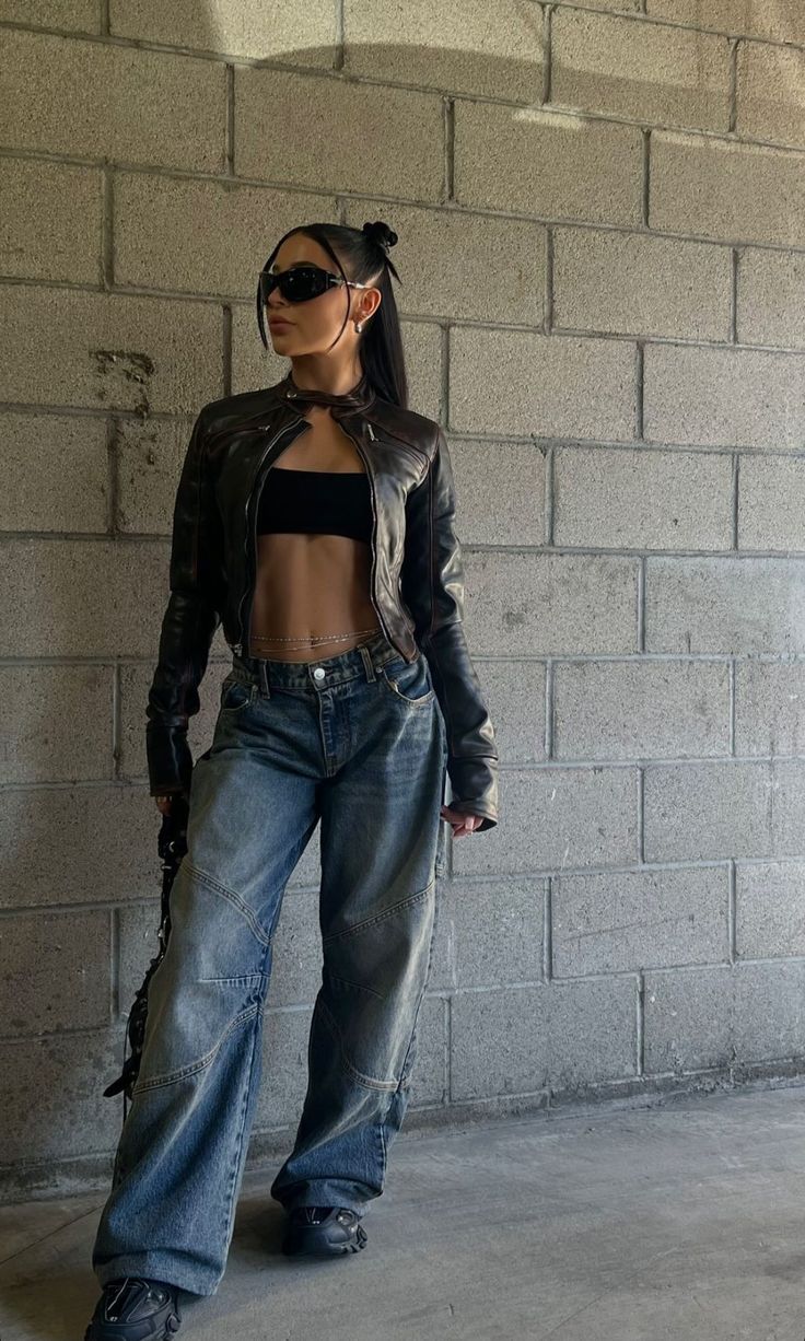 Woman in a leather jacket, crop top, and baggy jeans against a concrete wall