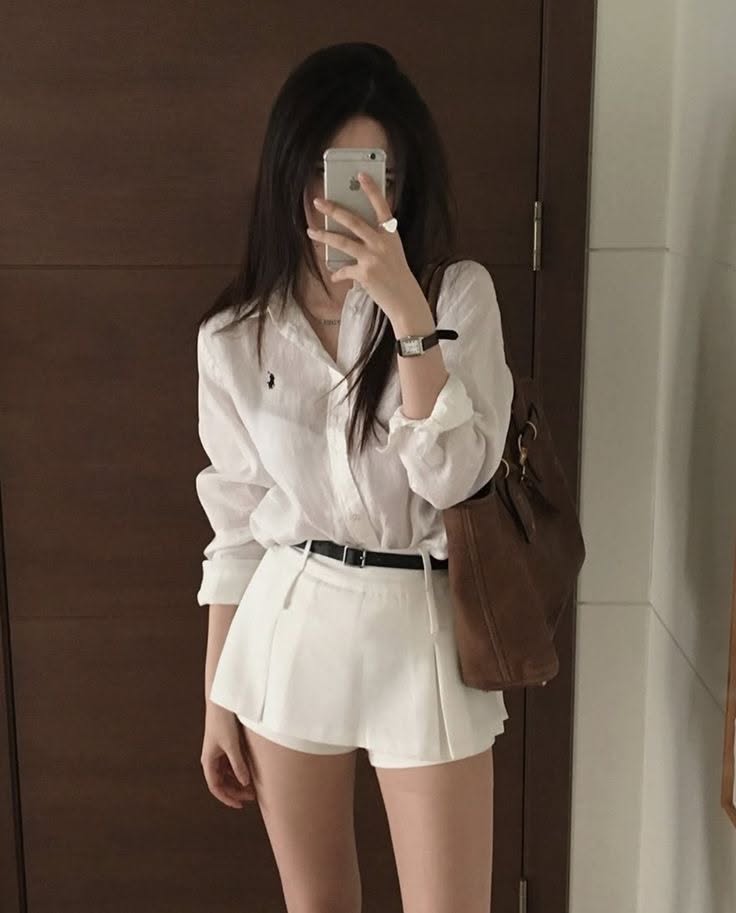 Woman in a white shirt and shorts taking a mirror selfie indoors