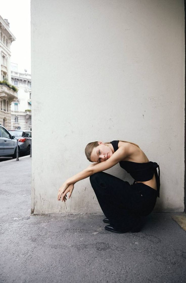 Woman in black top and pants squatting on a street next to a wall