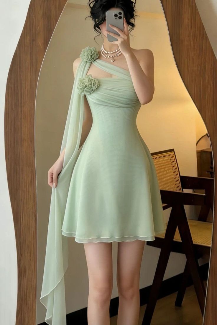 Woman in a light green one-shoulder dress with pearl necklace taking a mirror selfie