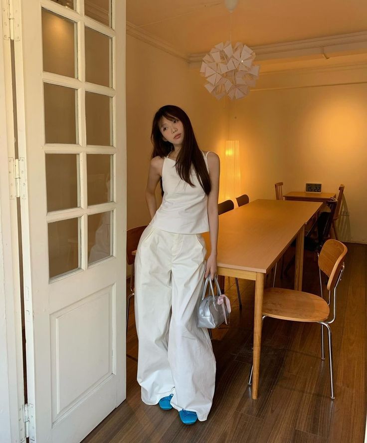 Woman in a white top and wide leg pants with blue shoes and a silver bag indoors