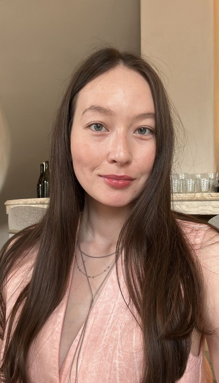 Woman with long brown hair wearing a pink top and layered necklaces indoors