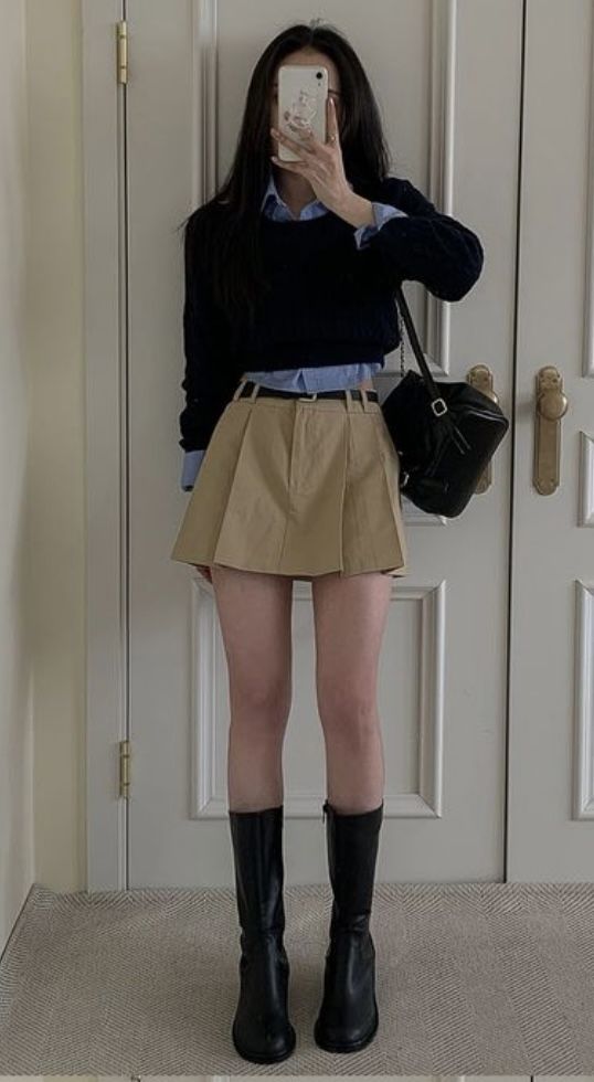 Person in a pleated skirt, long sleeve shirt, and black boots taking a mirror selfie