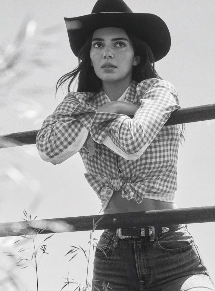 Woman in a cowboy hat and gingham shirt leaning on a fence outdoors
