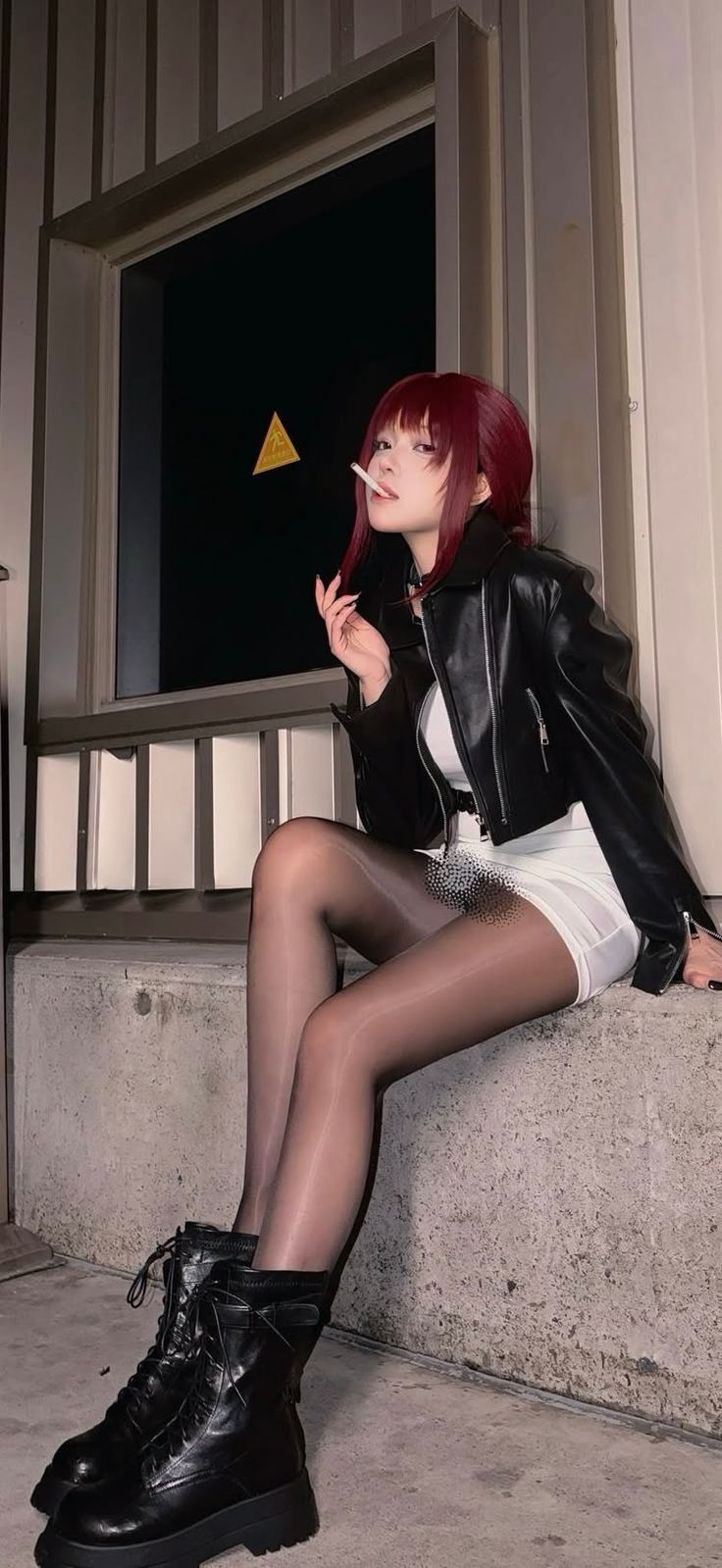 Woman with red hair in a leather jacket and white dress, smoking outdoors at night