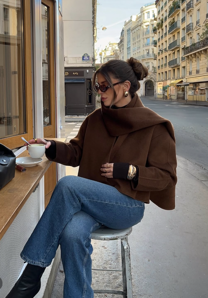 Woman in a brown jacket and blue jeans sitting outdoors on a street