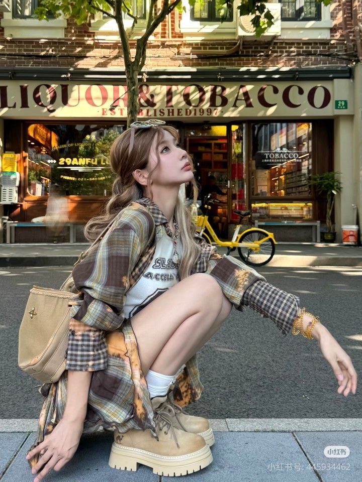 Woman in a plaid shirt and chunky boots crouching on a street in front of a liquor store