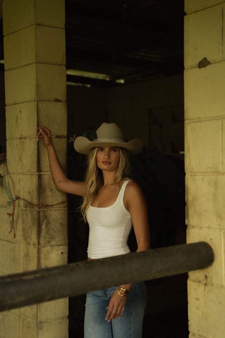 Woman in a cowboy hat and white tank top leaning against a pillar indoors