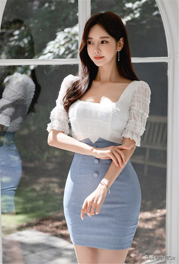 Woman with long hair in a white top and blue skirt standing by a window