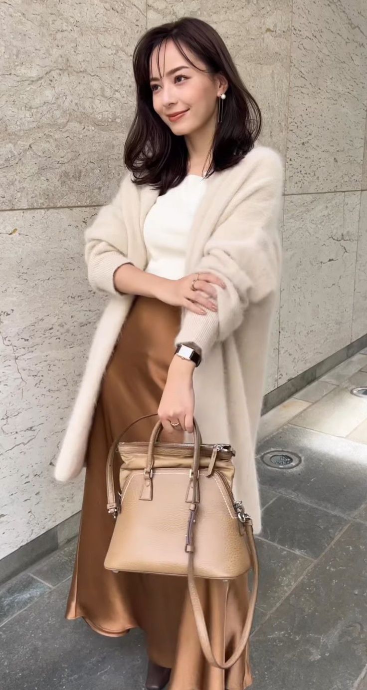 Woman in a long cardigan and brown skirt holding a handbag outdoors