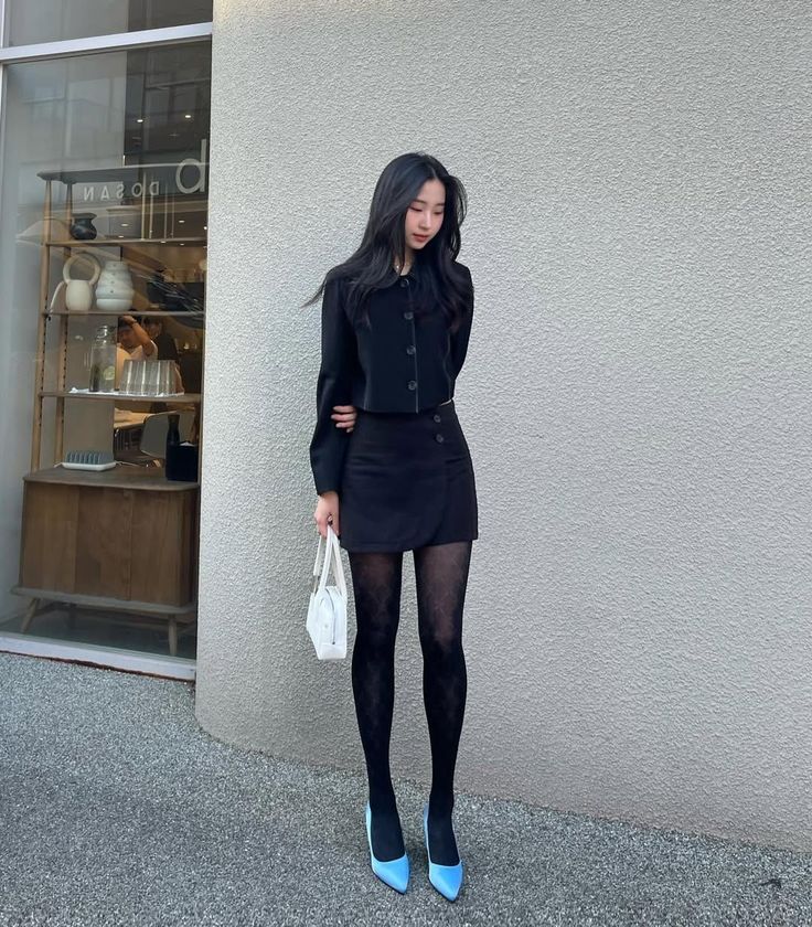 Woman in a black skirt suit and blue heels holding a white handbag outdoors