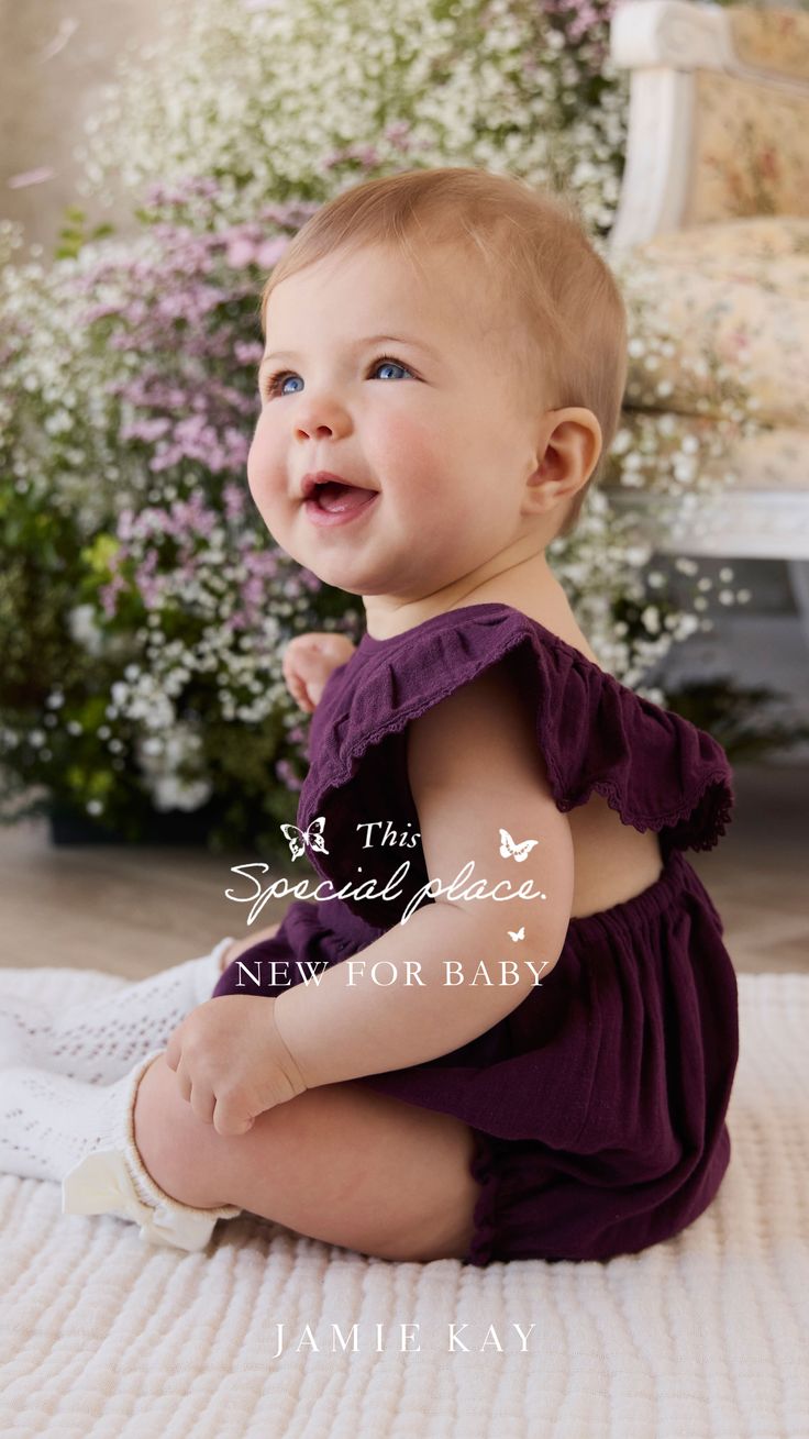 Happy baby in a purple romper sitting on a white blanket with flowers in the background