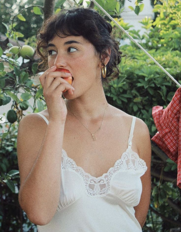 Woman in a white slip dress with lace trim eating an apple outdoors