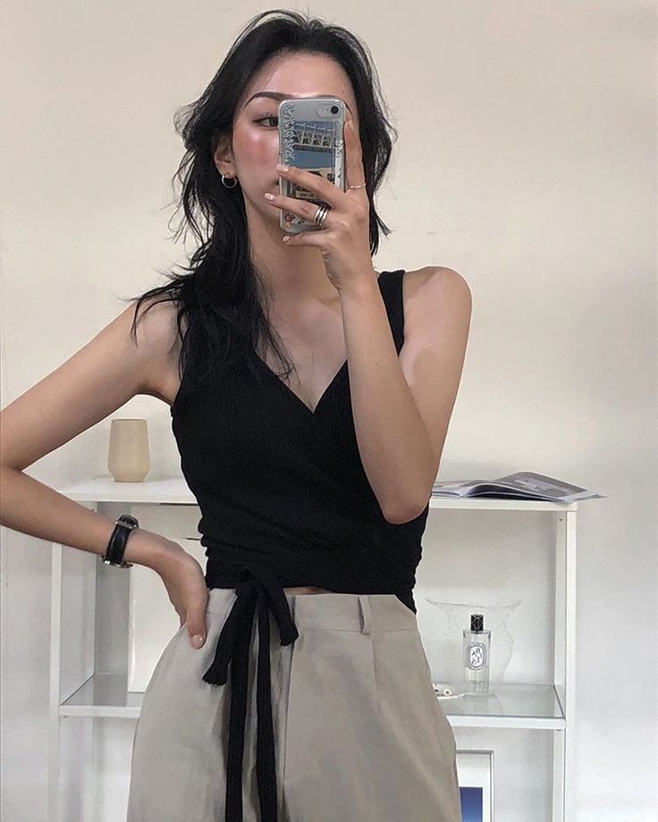 Woman in a black top and beige pants taking a mirror selfie indoors