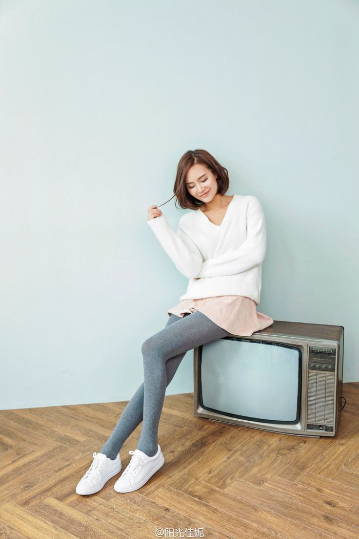 Woman with short hair in a white sweater and grey leggings sitting on a vintage TV